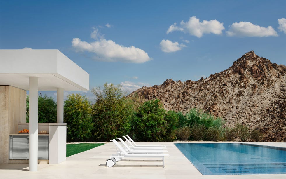 a covered outdoor kitchen is next to a zero edge pool with a large deck and four white lounge chairs that have back wheels, beyond the pool is grass, shrubs and trees, and a rocky mountain