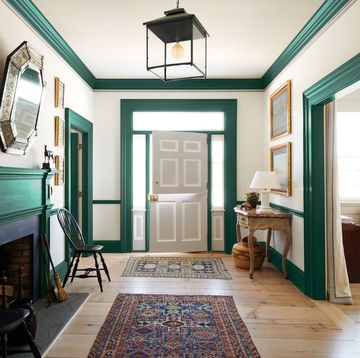 an entry hall has off white walls with rich green moldings and woodwork, a dutch door painted light gray, a fireplace, two black wooden chair, two caucasian rugs, a marble topped half moon console, and a gallery lantern