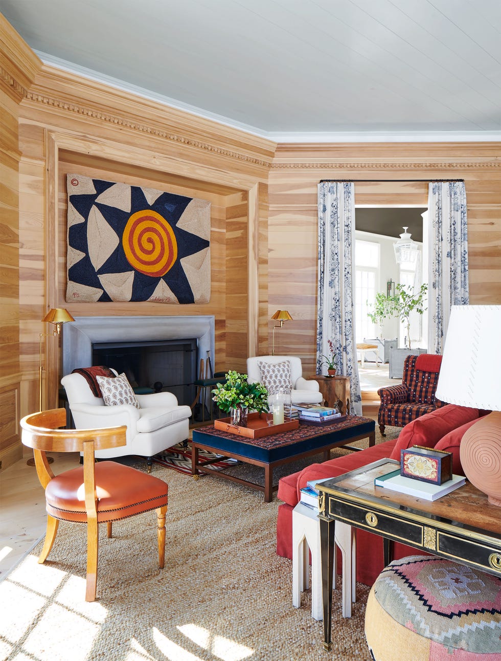 the walls of an octagonal room are horizontal pine boards, fireplace with large tapestry above, two white and one print upholstered chairs, fabric covered cocktail table, terra cotta sofa, printed curtains on doorway