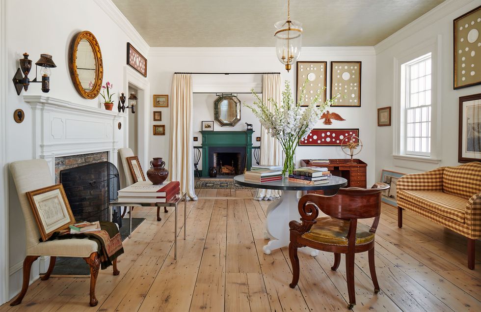 a white living room has a wide plank wood floor, a brick fireplace with sconces and mirror above, two armless chairs, a round pedestal table and antique armchair, checked sofa, a small desk, and multiple wall artworks