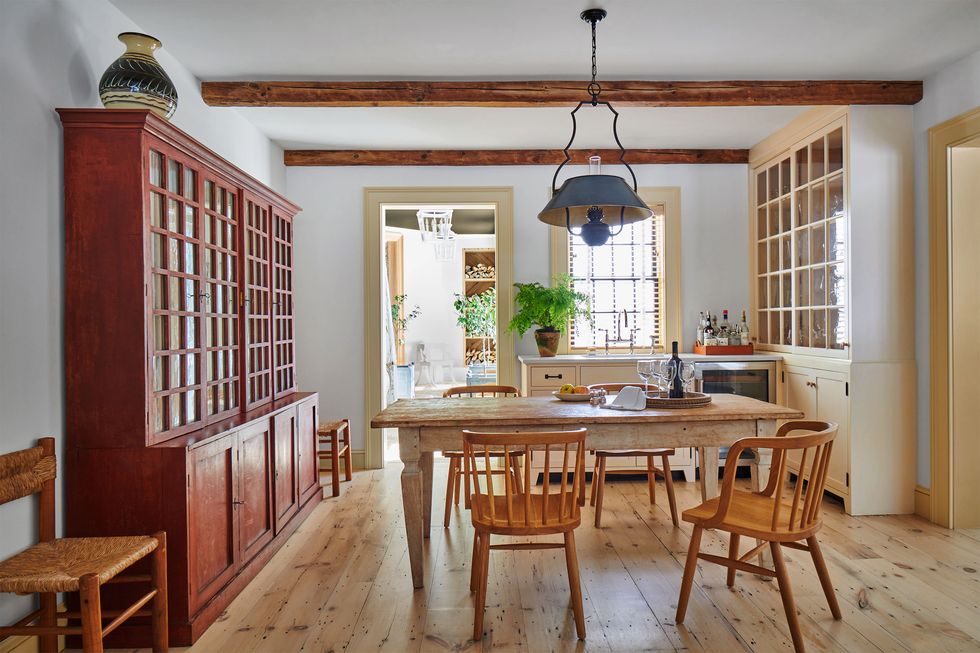 a kitchen has two large sideboards with cabinets below and glass windowpane storage above, a rustic wood table with four armchairs and two side chairs, a blue pendant over table, wood floors, and a beamed ceiling