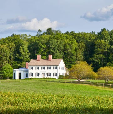 a colonial style white house with two floors and black shuttered windows with an octagonal single floor addition sits on a property with an expansive front lawn with a rustic horse fence, and tall trees behind the home