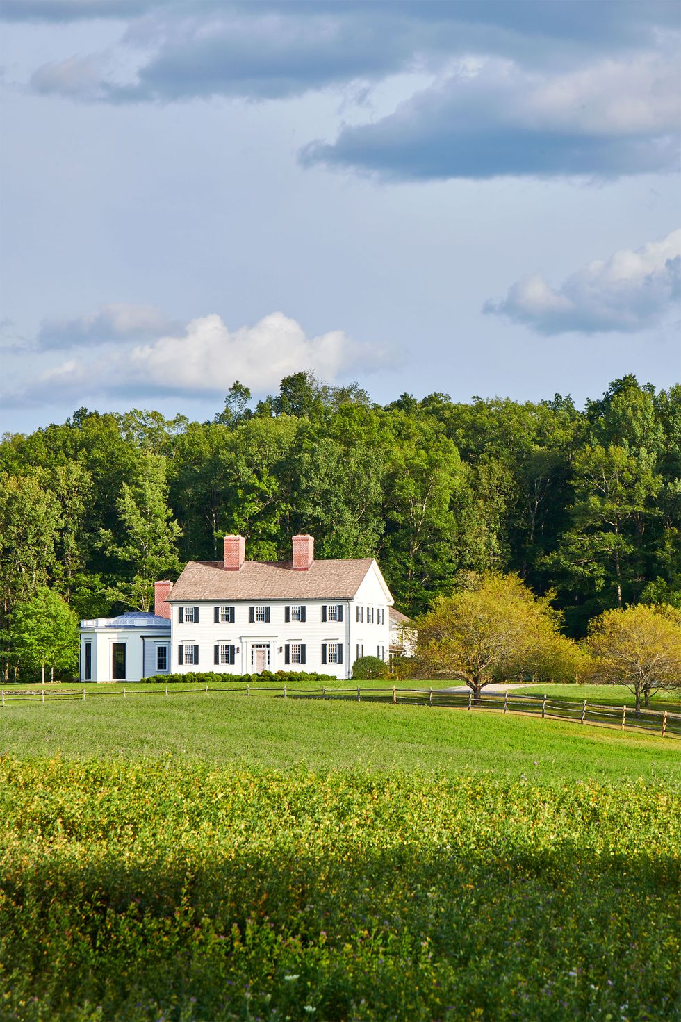 a colonial style white house with two floors and black shuttered windows with an octagonal single floor addition sits on a property with an expansive front lawn with a rustic horse fence, and tall trees behind the home