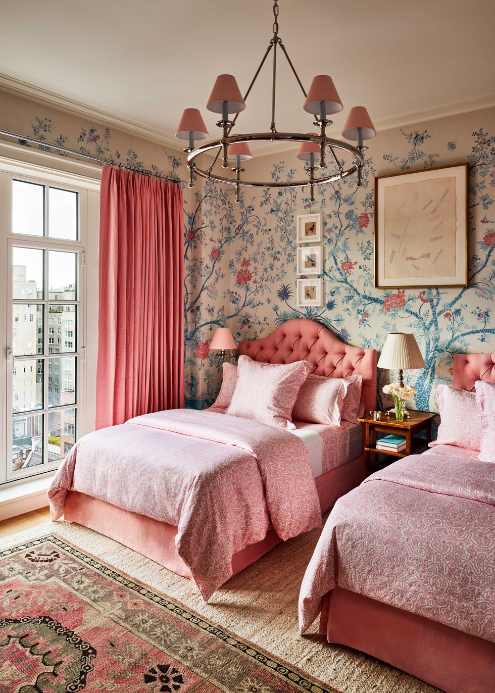 twin beds with a salmony pink upholstered headboards and matching curtains, linens in a patterned pink fabric, floral and tree print wallpaper, wooden nightstands with lamps, chandelier with six small shades in matching pink