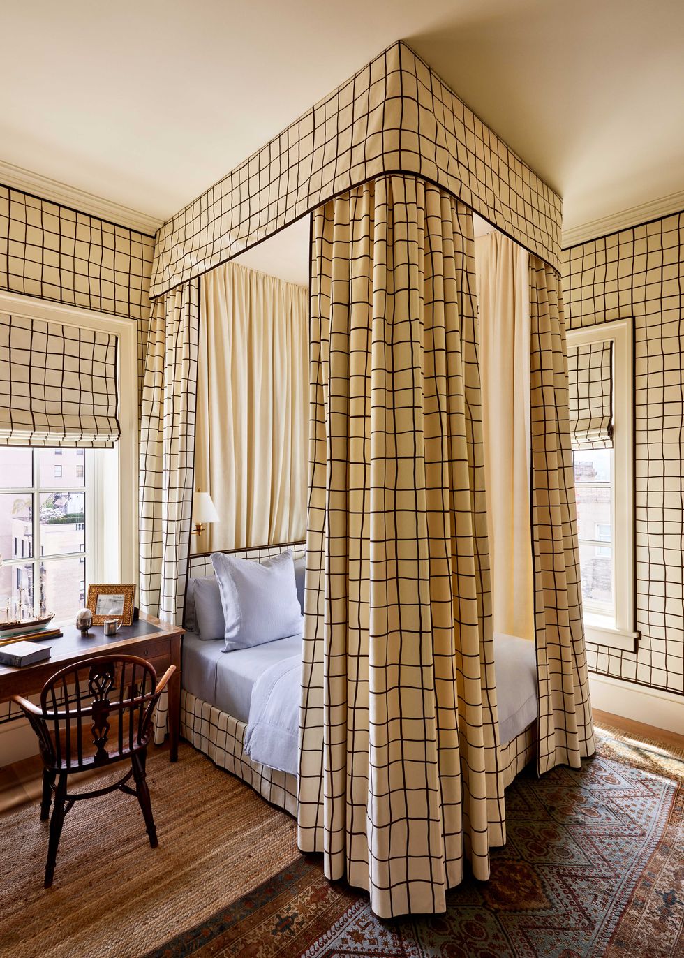 a bed with light blue linens is surrounded by a canopy and curtains in a light yellow with black windowpanes fabric, which matches the wallpaper and window shades, a wooden writing table and chair sit next to the bed
