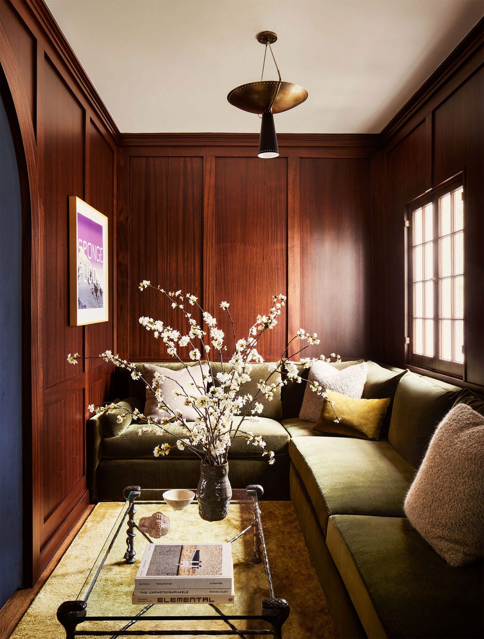 a wood-paneled dining room corner with a light green sofa and a glass-topped cocktail table with a vase holding beautiful branches