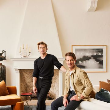 in a white living room with blue carpet, a white sofa, and fireplace are two men, one standing wearing a black sweater and pants, the other seated on the sofa back wearing a tan jacket, white shirt, and black jeans