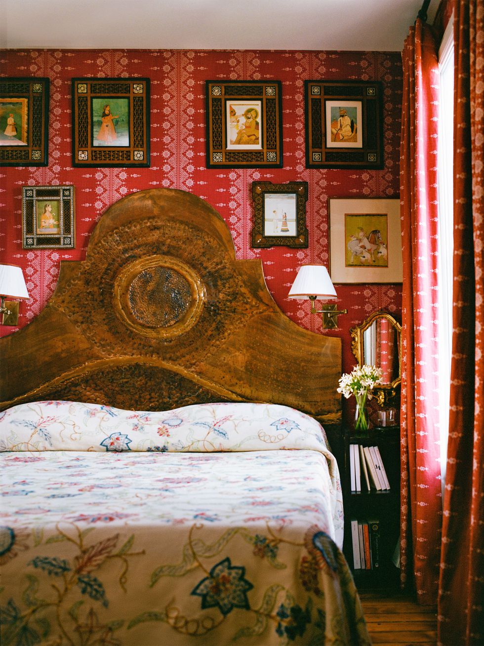 a large bed with a hammered copper headboard flanked by sconces with shades, floral printed bedcover, small bookshelf with mirror above, multiple framed artworks against red and white patterned wallpaper and curtains