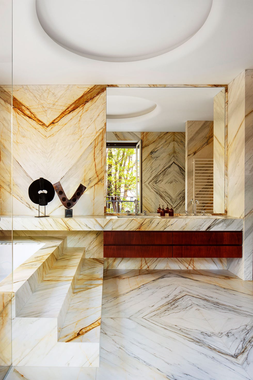 a bathroom with a white and ocher marble floor and three steps up to a bathtub, a counter with two sinks, two meal sculptures, wood cabinets beneath and large mirror above counter