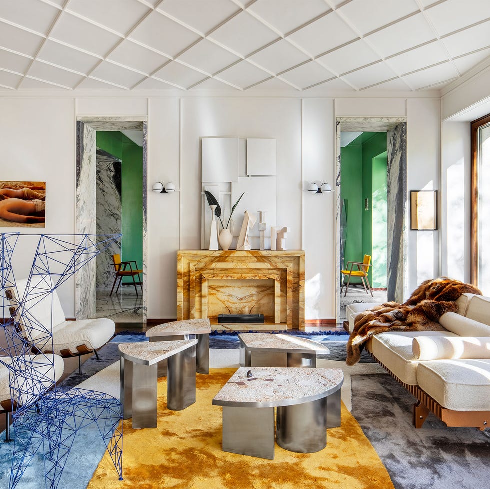 Hannes Peer - ELLE DECOR living room with white walls and ceiling with square panels, ocher colored marble fireplace, blue and ocher rug, white boucle chairs and sofa, blue wire chair, metal based cocktail tables with marble tops