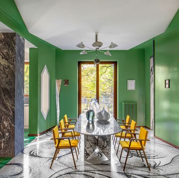 hexagonal dining room with green walls, oblong white and black marble table, wood chairs with orange fabric seats, glass sculptures on table, five armed pendant, glass floor lamp, patterned floor