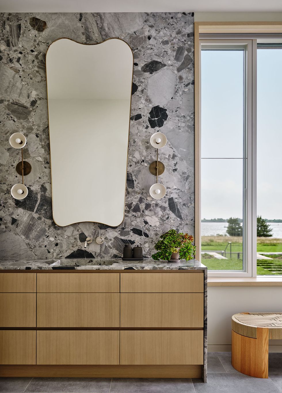 a bathroom with a marble wall with a irregular shaped mirror flanked by sconces, wood cabinets with marble top and sink, potted pant, circular two toned bench, windows looking out to lawn and the sea