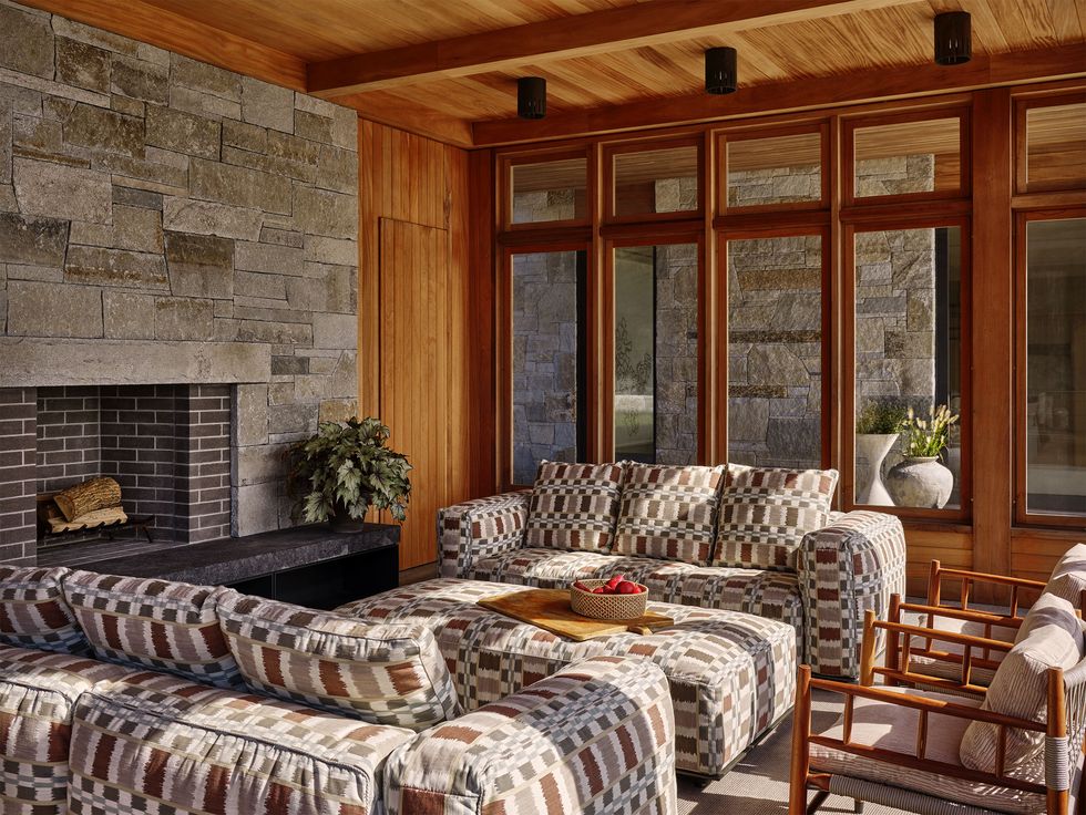 on a screened in porch are two sofas and an ottoman with a small geometric pattern in brown, gray, and white, two wood chairs with fabric cushions, a brick fireplace, wood ceiling and frames for large windows