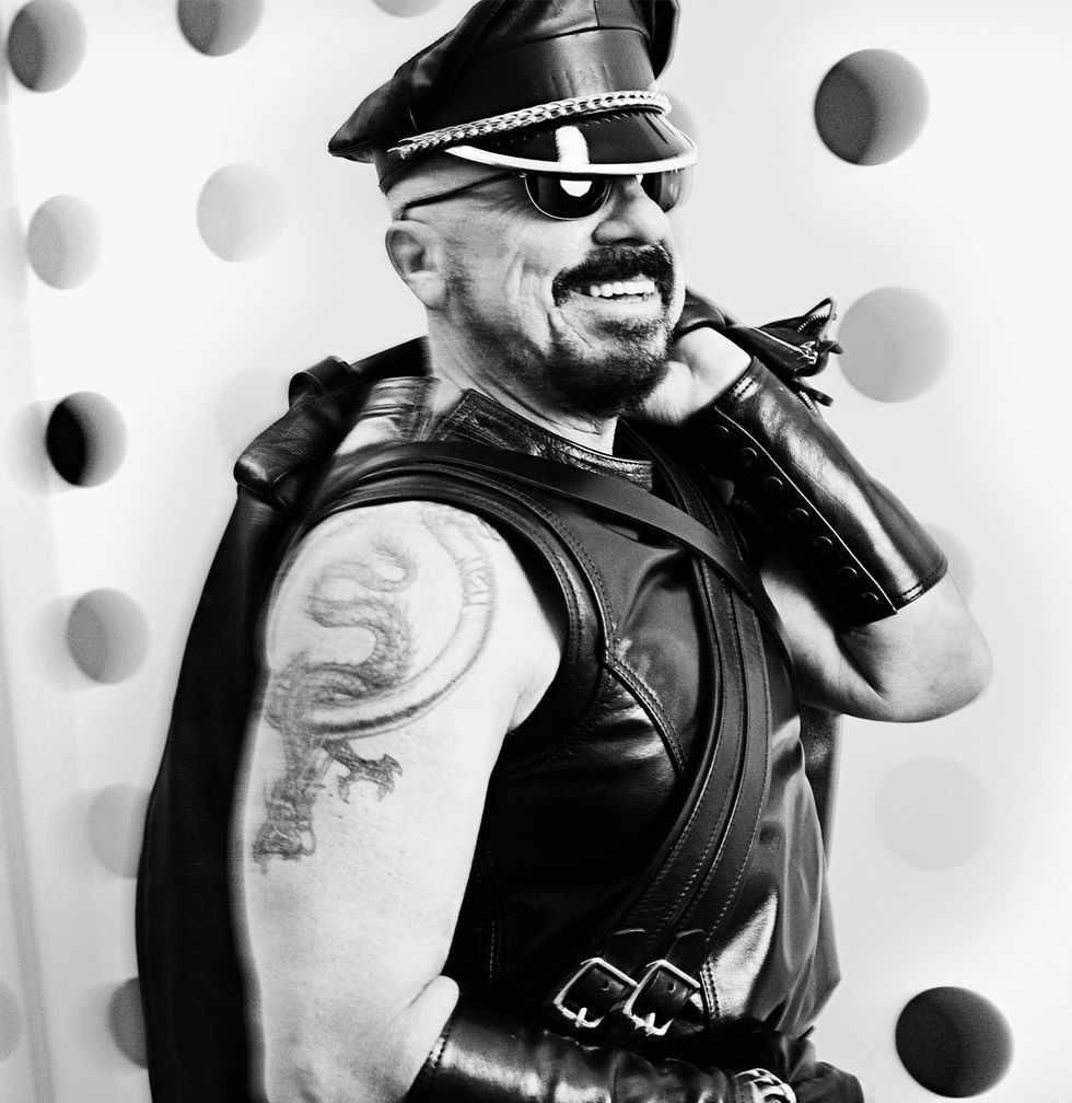a bearded man photographed at a side angle from the waist up, dressed in sleeveless leather attire with leather wrist and forearm guards, a dragon tattoo on his right upper arm, a leather hat, and sunglasses