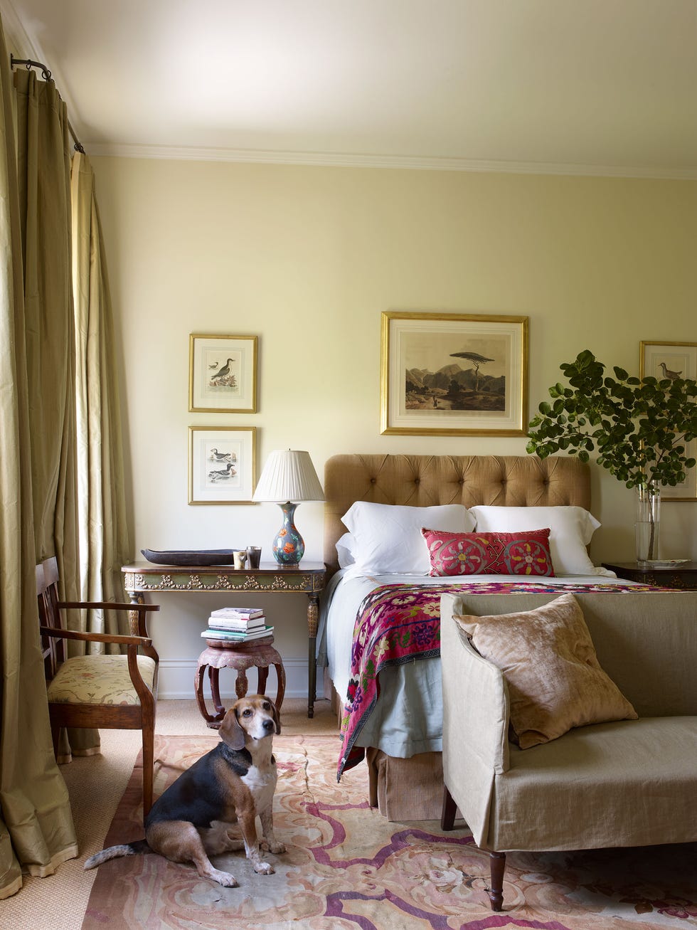 Julia Reed - ELLE DECOR bedroom with tufted headboard on bed and artwork on the wall and a long side table and large settee at the foot and the most