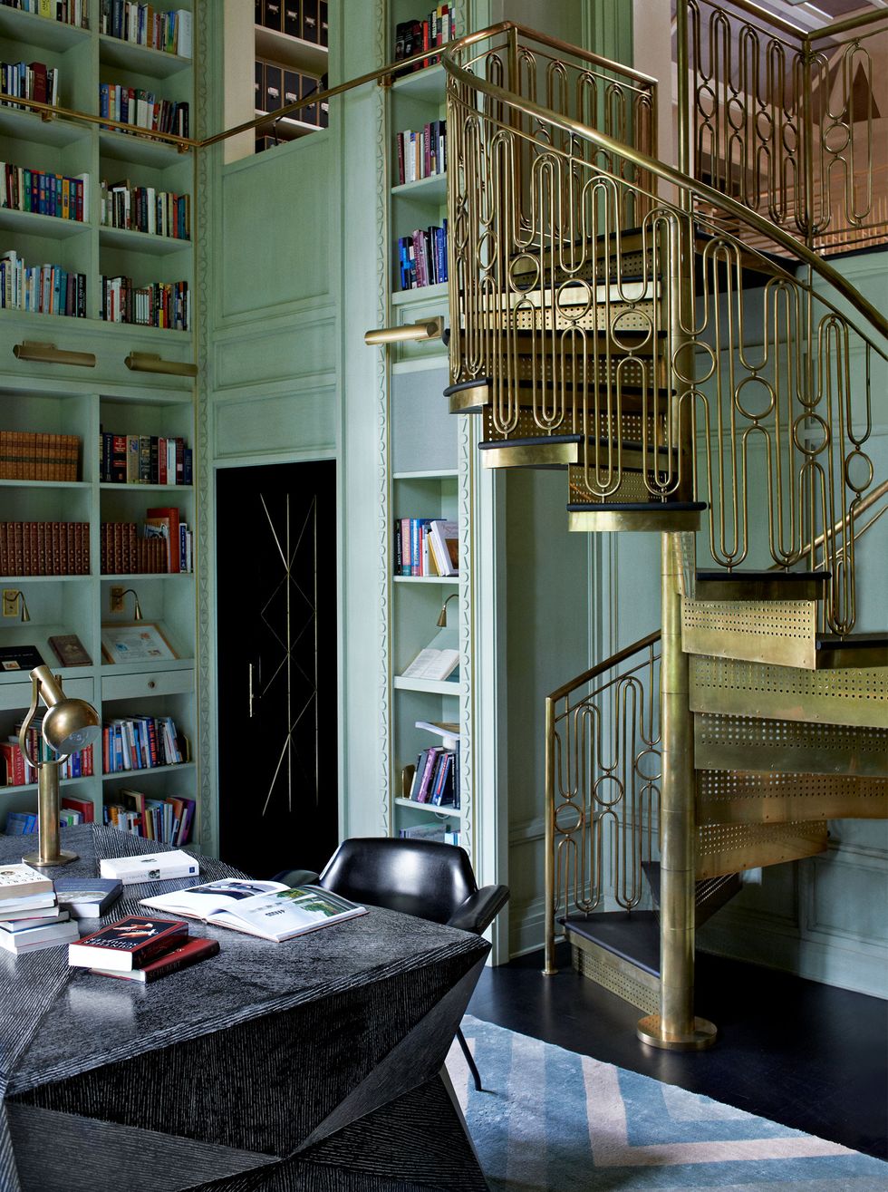small brass art deco spiral staircase in a library with light colored floor to ceiling shelving and a marble desk and leather chair and blue patterned geometric rug