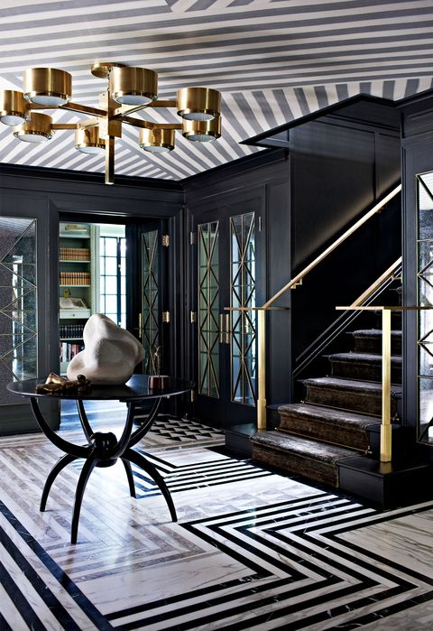 glamorous black entry hall with dark steps and bronze banisters leading up and a black table at center with spider like legs and a floor with zigzag lines in various tones and a set of doors next to the staircase