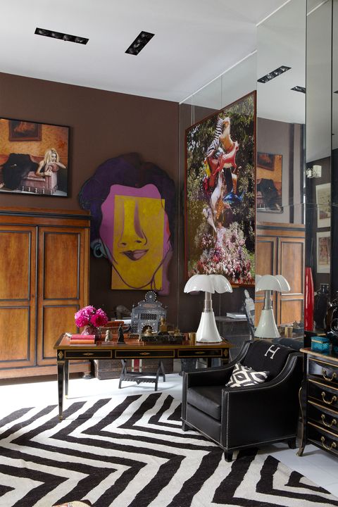 corner detail with black and white stripey rug and black leather chair and black inlaid desk with a white lamp and a wood commode at back and a large colorful face painting on the wall and other pieces of art