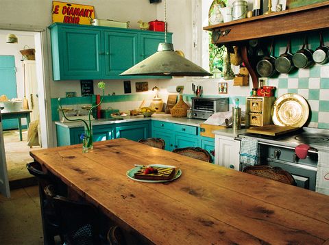 in the kitchen is a rustic wooden table with caned back antique chairs and a pendant above, turquoise cabinets, a stove with green and white tiles on the wall behind it, and pots hanging from a wooden shelf