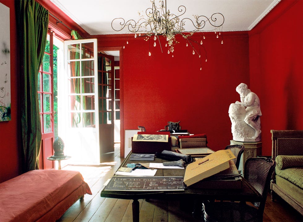 a sunlit door with green drapes leads to a red music room, a long table with books and papers, antique chairs and daybed, a statue of a young michelangelo, and a chandelier sculpture with glass beads