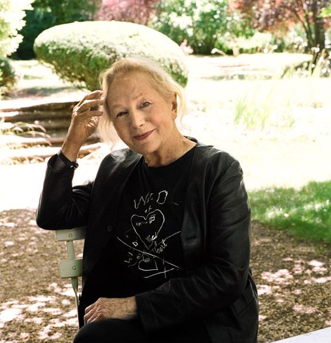the designer sits in a bistro chair in her garden, wearing a light black jacket over a black t shirt with a white abstract design