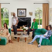mara brock akil wearing a long discreetly sheer applique dress with strappy sandals and seated on a green sofa and the husband standing behind and two grown sons seated on an opposite matching sofa with a small coffee table between them  in a room with long cream colored curtains and two doors that open to an outside garden