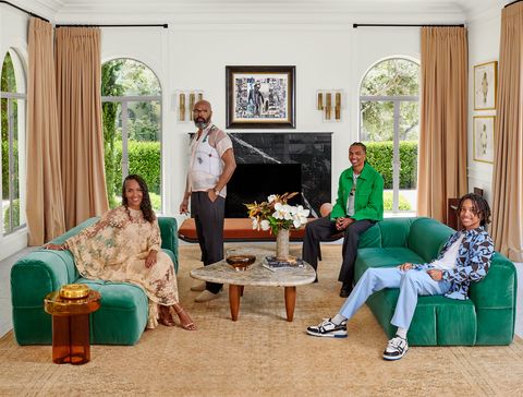 mara brock akil wearing a long discreetly sheer applique dress with strappy sandals and seated on a green sofa and the husband standing behind and two grown sons seated on an opposite matching sofa with a small coffee table between them in a room with long cream colored curtains and two doors that open to an outside garden