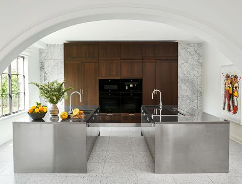 a kitchen has two facing steel islands, both with a sink and cabinets below, appliances embedded in a wood cabinet against the back marble wall, windows on left and artwork at right, all viewed through an archway