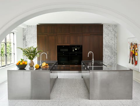 a kitchen has two facing steel islands, both with a sink and cabinets below, appliances embedded in a wood cabinet against the back marble wall, windows on left and artwork at right, all viewed through an archway