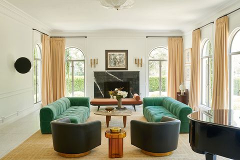 living room with surrounding doors with ceiling to floor cream colored curtains each leading to outside and with twin facing vintage italian sofas in green velvet  a cocktail table between them  and two rounded leather chairs will gold stands flanking each sofa
