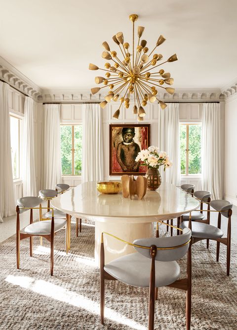oblong dining table in a delicious stone colored parchment with delicate vintage chairs in gold frames and white seats and back and a starburst chandelier, a dramatic painting of a child in a gilded frame, windows on all sides with long flowy white curtains