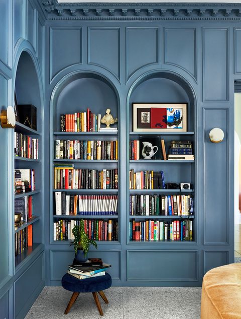 a library has blue painted built in shelves lining two walls filled with books and objets d'art, a short stool with wooden legs and a deep blue velvet seat, and a curved sofa in a plush light brown fabric