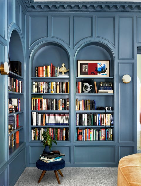 a library has blue painted built in shelves lining two walls filled with books and objets d'art, a short stool with wooden legs and a deep blue velvet seat, and a curved sofa in a plush light brown fabric