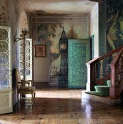 very dramatically lit entrance with a scroll like grandfather clock against the far wall and a scenic wallpaper behind it and in the foreground is a lone chair to the left and a very sweeping staircase with green runner to the right