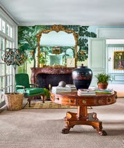an entryway has a fireplace with a decorative marble surround and ornate mirror above, wallpaper with floor to ceiling trees, a round polished wood pedestal table with a vase and books, and a green cushioned armchair
