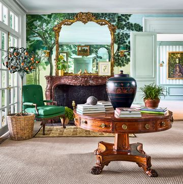 an entryway has a fireplace with a decorative marble surround and ornate mirror above, wallpaper with floor to ceiling trees, a round polished wood pedestal table with a vase and books, and a green cushioned armchair