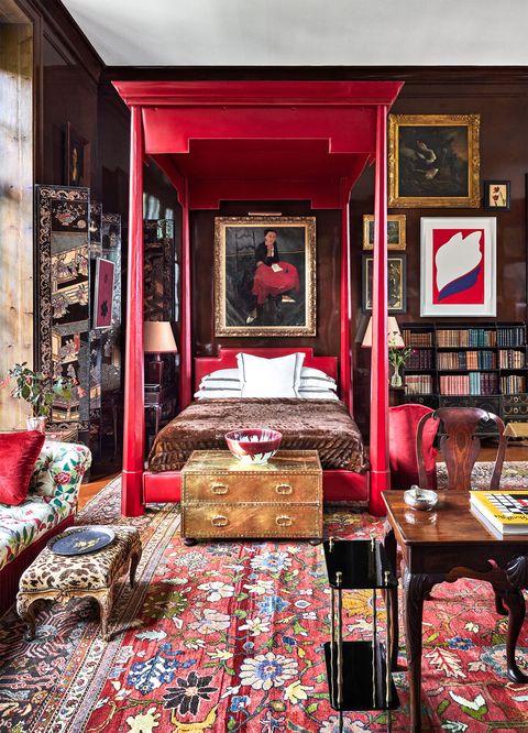 a living room also has a red based floral rug, a floral print sofa and animal printed ottoman, a wooden writing table and chair, a red leather canopy bed with a portrait hanging above it, other artworks and book shelves