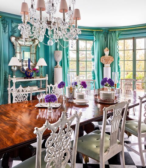 a dining room has turquoise walls and curtains, a long wooden pedestal table, chairs with white filigreed backs and green fabric seats, black and white square tiled floor, a crystal chandelier, and a bust sculpture on a plinth
