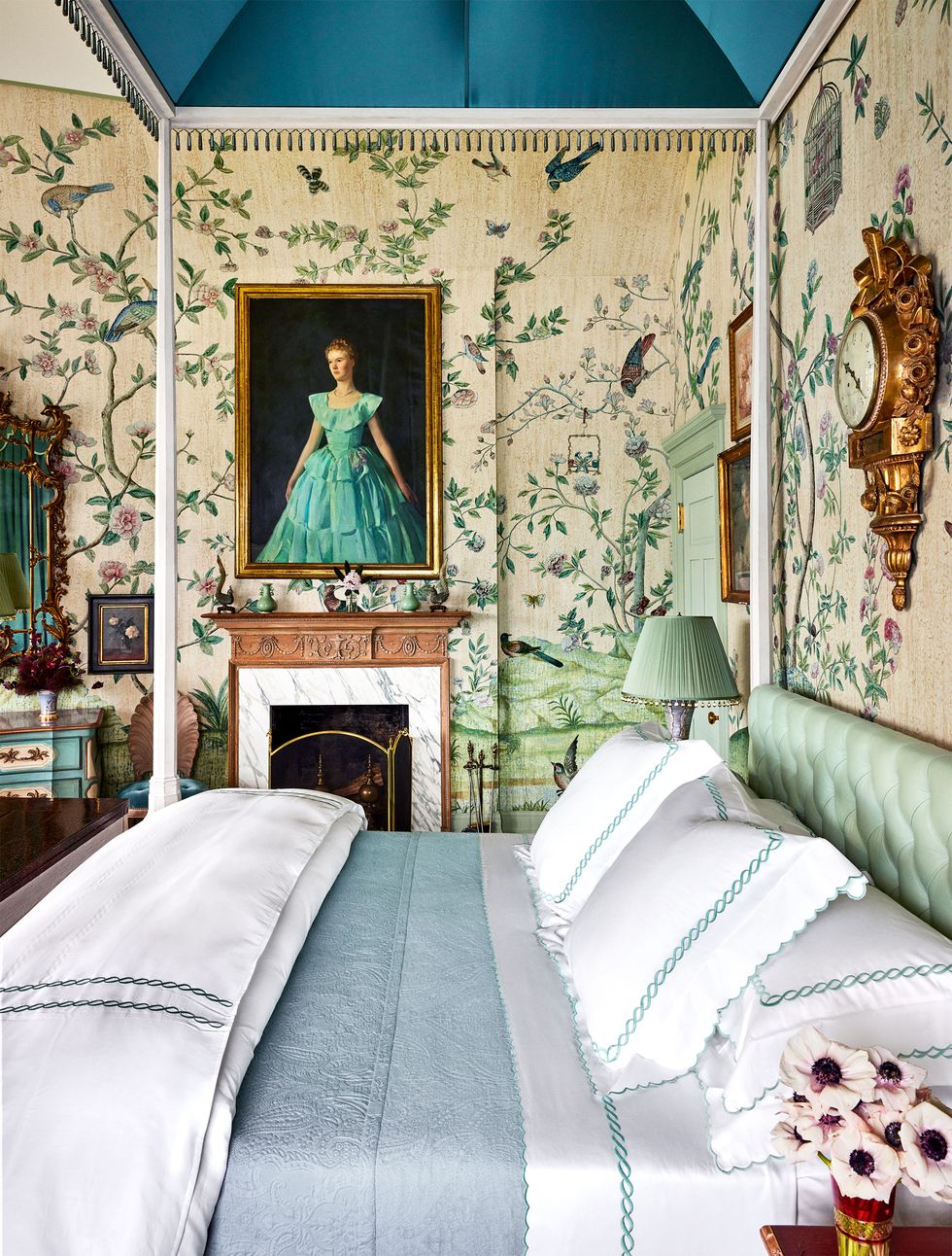 The bedroom has vine floral wallpaper with birds on it, a small fireplace, above which is a portrait of a young girl in a green skirt, and the bed has a tufted light green headboard. There is a matching pleated lamp next to it, with starched white linen.The bed has a twisted rope design and a light blue comforter.