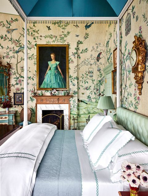 bedroom with a viney floral pattern wallpaper with some birds on it and a small fireplace and a portrait of a young girl in a green skirt above it and the bed has a tufted light green headboard with a matching pleated lamp next to it and starched white linens on the bed with a rope twist design along them and a light blue coverlet