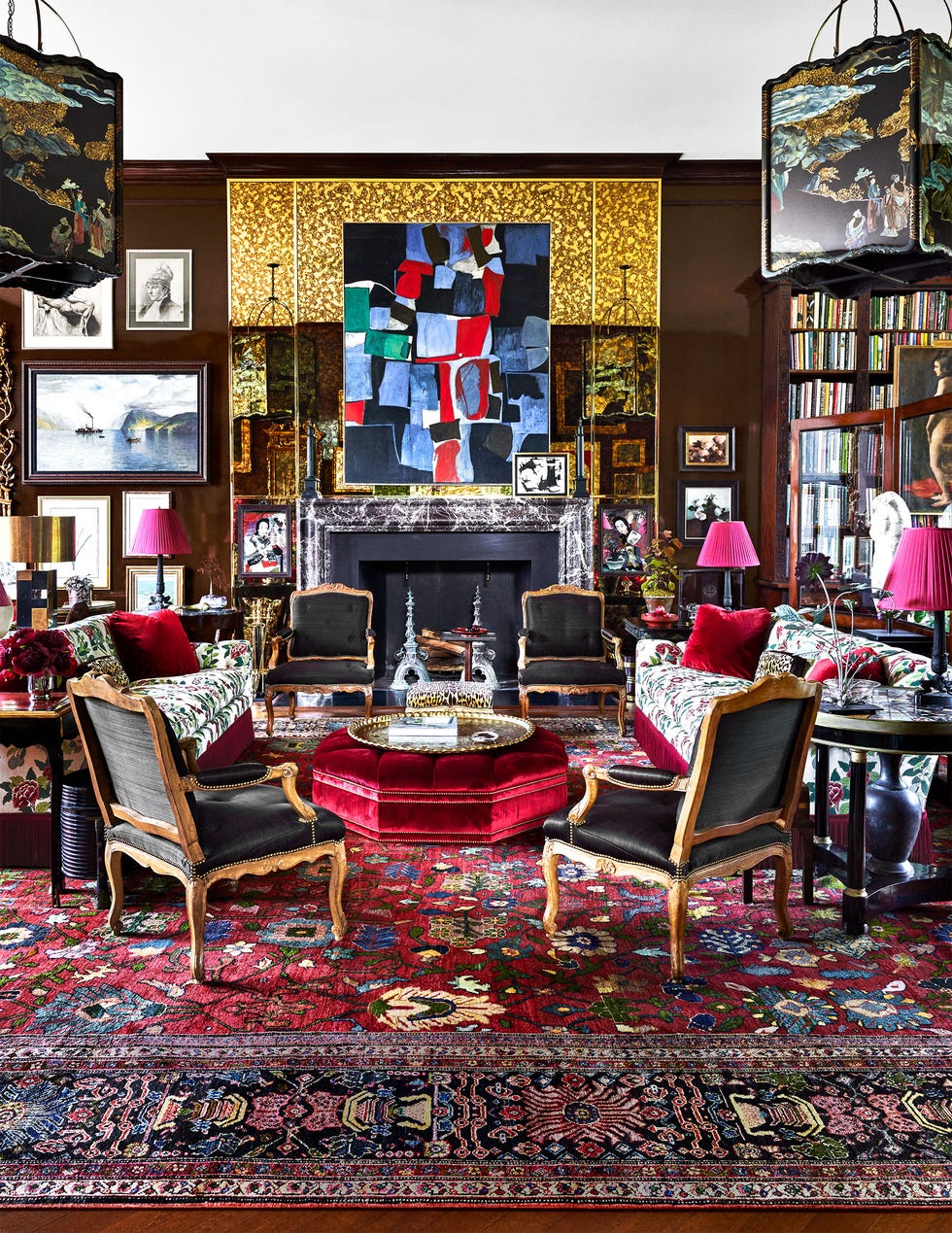 in a living room is a black fireplace with a marble surround and an abstract artwork above, multiple other artworks on the walls, two flower print facing sofas, four armchairs with black cushions, and a red velvet ottoman