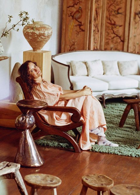 elena reygadas wearing a long flowly peachy dress and seated comfortably in a wooden sloop back chair as she holds one knee up and looks at the camera and the chair is on both the wooden floor and the green knotted rug and there some small stools around and some artwork
