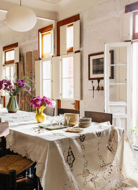 long rustic table with cotton tablecloth with lace cutouts and a few colorful jugs and stacked plates and rush chairs pulled up to it in a whitewashed room with lots of windows and doors for light