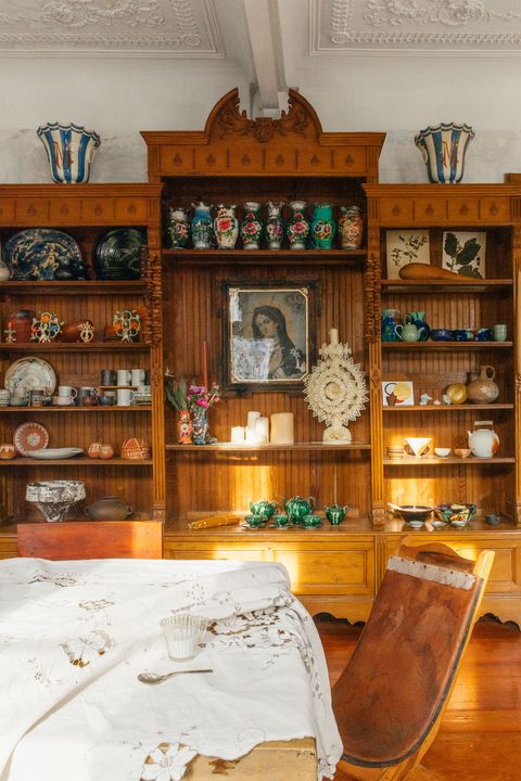 wall size built in shelves with pottery and small scultpure and religious painting right at center and in the foreground is a cotton lacework tablecloth over a rustic table and a wooden scoop chair pulled up to it