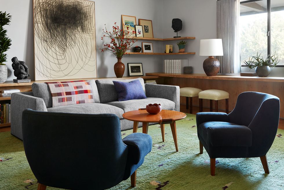in a den are two vintage armchairs in a deep blue fabric, a sofa in a light gray fabric, wood cocktail table, a wool rug in a light green hue, multiple artworks and a lamp on shelves and on a counter bar