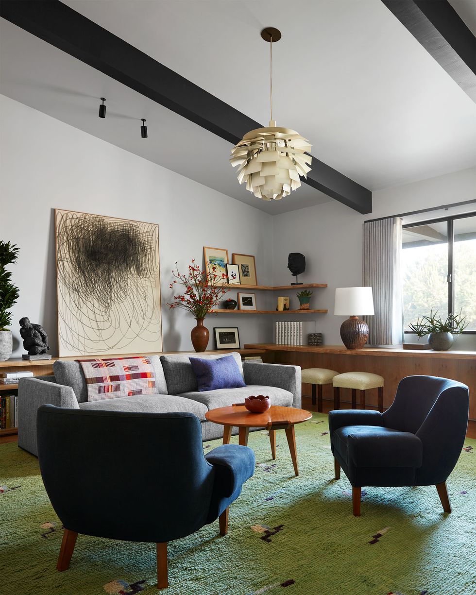 in a den are two vintage armchairs in a deep blue fabric, a sofa in a light gray fabric, wood cocktail table, a wool rug in a light green hue, multiple artworks and a lamp on shelves and on a counter bar