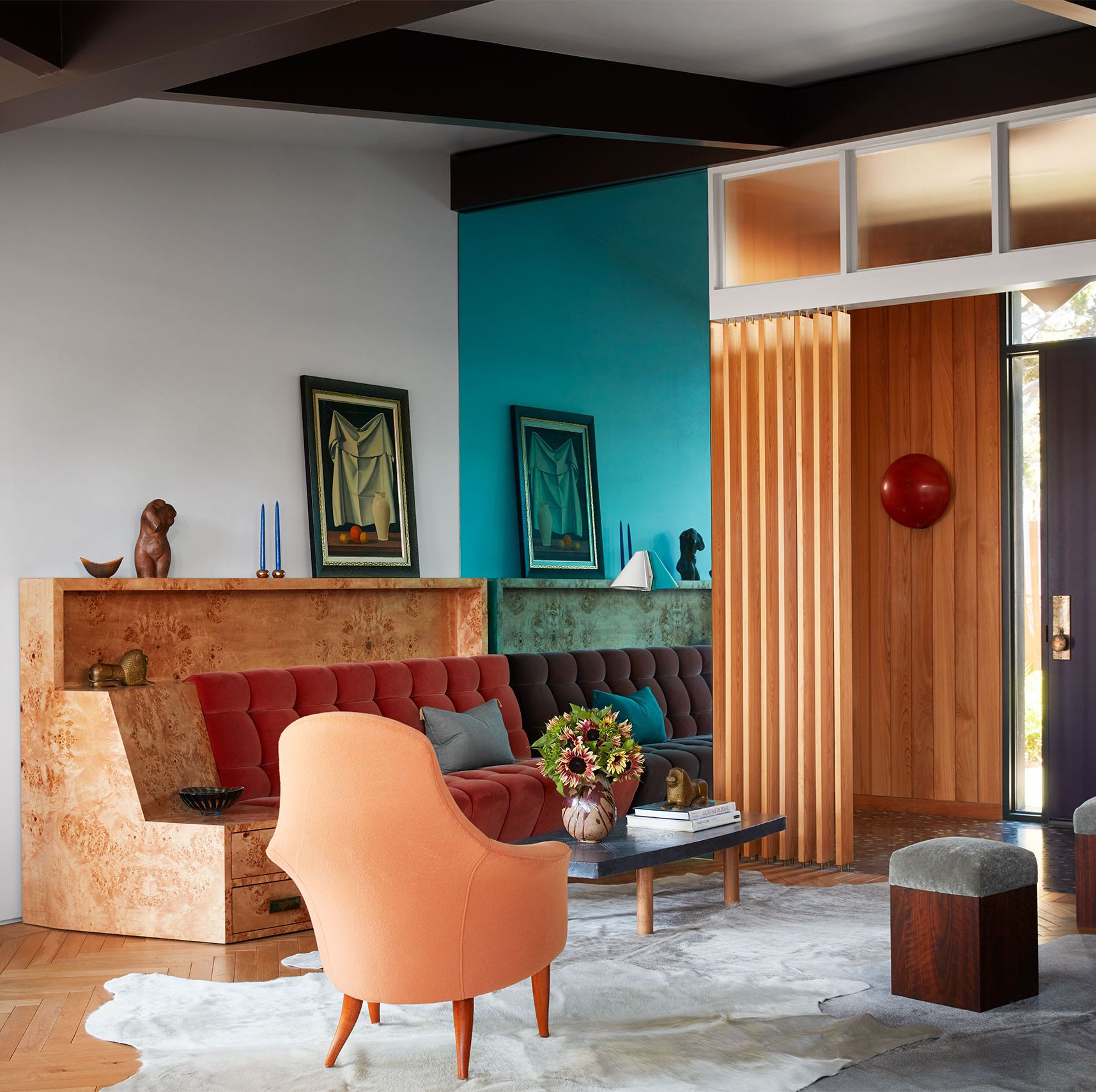 in a midcentury beach house living room is a bar  fronted with colored tiles, a sofa with a burl wood frame and tufted terra cotta cushions, two modern chairs, cocktail table, too stools, and a cow hide rug
