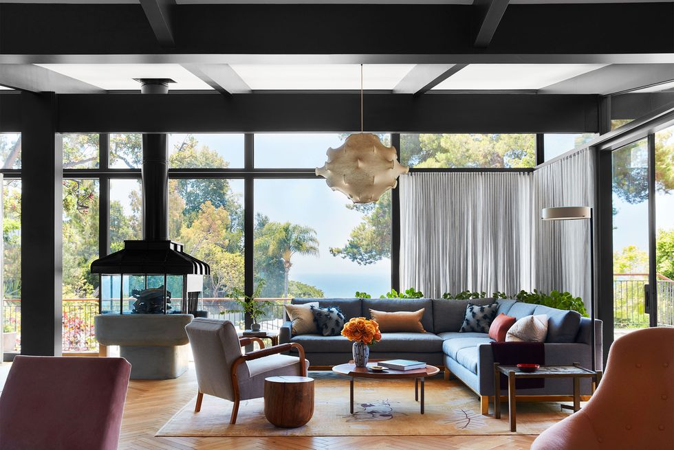 a living room with floor to ceiling windows, some with curtains, a pewter colored fabric sectional, an upholstered chair, cocktail and end tables, a light wool rug, a cocoon pendant, and an indoor fire pit