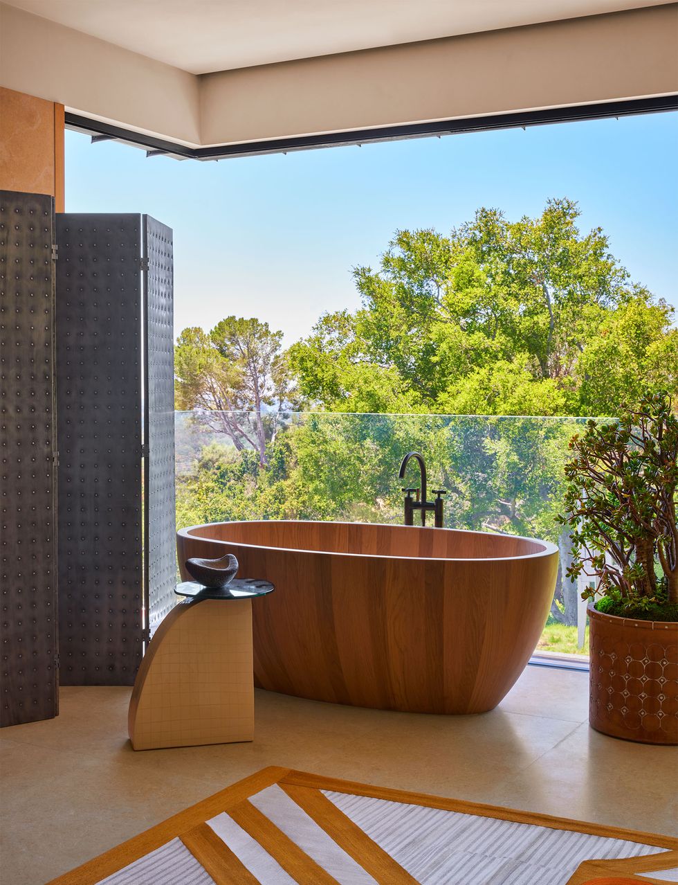 in a primary bathroom with a glass bottom wall and open top is an aluminum folding screen, a ceramic side table next to an oak barrel bathtub, a large ceramic planter, and a light rug with terracotta stripes