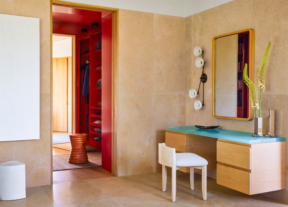 in a corner of a bathroom with limestone and plaster walls is a floating white oak vanity with glass top, a stool with a half back, a wall mirror and a sconce with four lights, to the side is a dressing room painted red